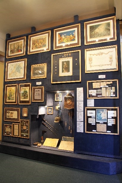 Auf dem Bild ist eine Ausstellungswand zu sehen, die sich mit einem Kriegsgefangenenlager in Krasnojarsk befasst. Sie ist an einer großen, dunkelblauen Wand installiert. An dieser Wand hängen diverse gerahmte Dokumente, Zeichnungen, Fotografien und Karten, die sich thematisch auf das Lager beziehen.