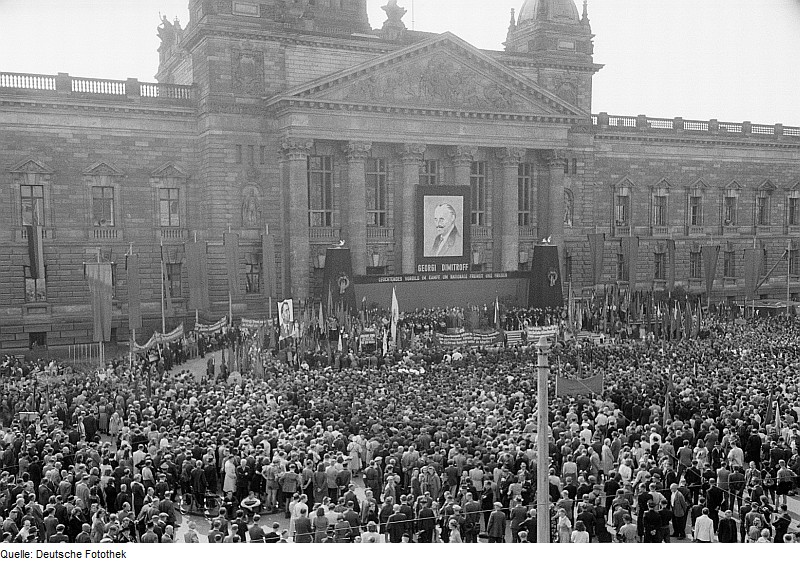Eine historische Schwarz-Weiß-Aufnahme zeigt eine große Menschenmenge vor einem monumentalen Gebäude mit klassizistischer Architektur. Das Gebäude verfügt über eine breite Fassade mit hohen Säulen und einem Dreiecksgiebel. Über der Menge hängt ein großes Porträt eines Mannes, flankiert von der Aufschrift „GEORGI DIMITROFF“. Die Menschen stehen dicht gedrängt auf einem Platz. Viele von ihnen halten Fahnen, Plakate und Transparente hoch. Die Atmosphäre wirkt wie bei einer politischen Kundgebung oder Demonstration.