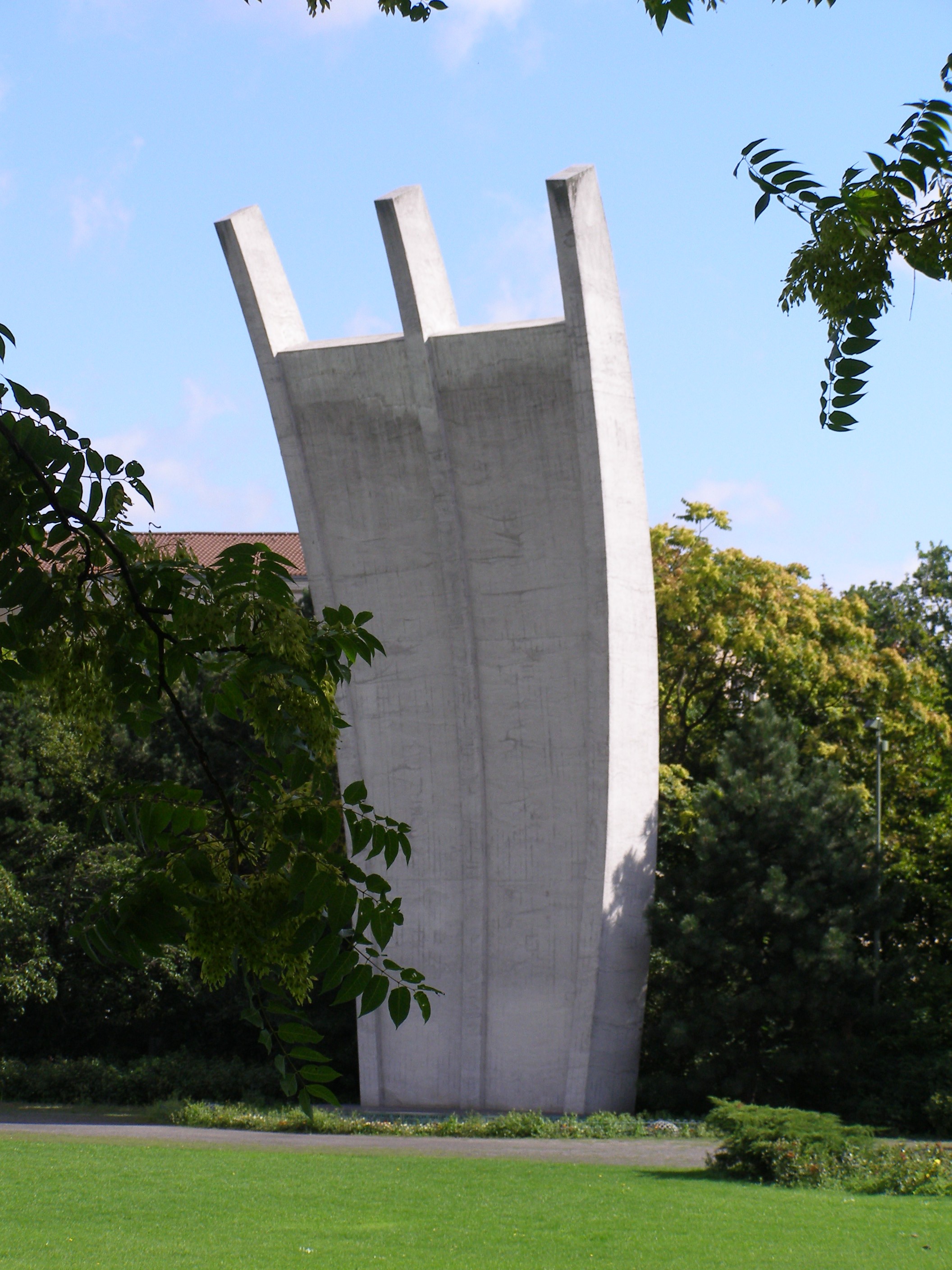 Ein hohes, schmales Denkmal aus Beton steht in einer grünen Parkanlage. Das Denkmal besteht aus drei senkrechten, leicht geneigten Platten, die nach oben hin auseinanderlaufen. Im Hintergrund sind Bäume und Sträucher zu sehen, und der Himmel darüber ist klar und blau. Der Rasen um das Denkmal herum ist gepflegt.