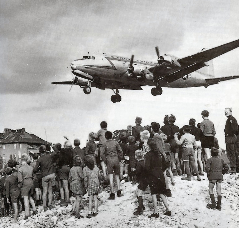 Das historische Schwarz-Weiß-Foto zeigt eine Gruppe von Kindern und Jugendlichen, die auf einem sandigen oder schuttbedeckten Hügel stehen. Sie beobachten ein großes Propellerflugzeug, das tief fliegt und sich im Landeanflug befindet. Im Hintergrund sind einfache Gebäude und ein bewölkter Himmel zu erkennen. Die Szene wirkt lebhaft.