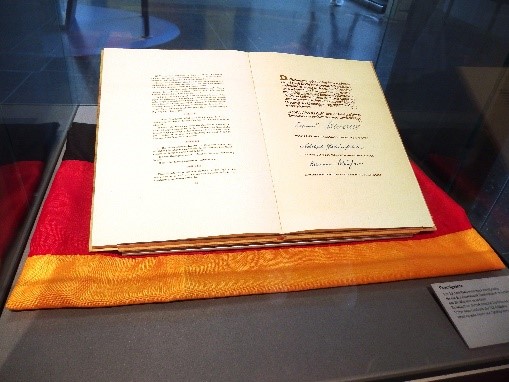 Das Bild zeigt ein aufgeschlagenes, historisches Buch, das in einer Vitrine auf einem schwarz-rot-goldenen Tuch liegt. In den Seiten des Buches sind handschriftliche Einträge und Unterschriften zu sehen. Rechts neben dem Buch ist eine kleine Beschriftungstafel angebracht.